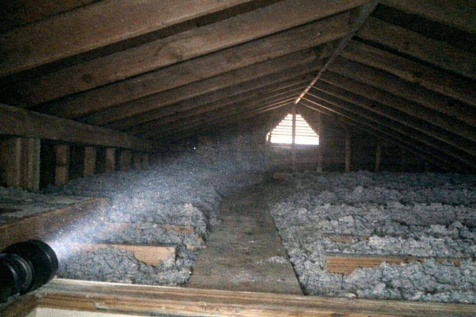 A flashlight beam raking across the rafters and blown-in insulation of a low attic toward a gable vent
