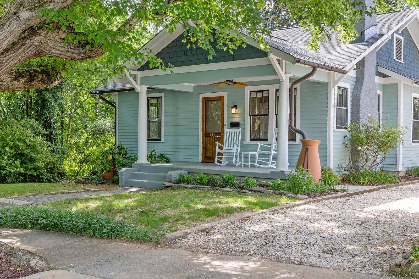 A blue Craftsman bungalow with a deep front porch and rocking chairs under mature trees