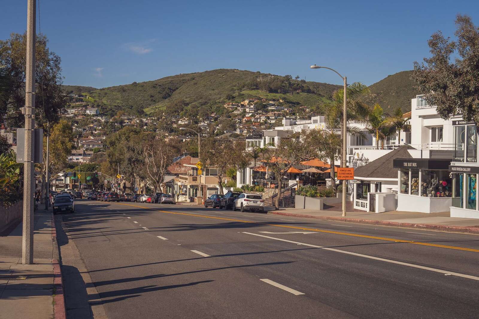A sunlit street of stucco buildings with dry hills rising behind